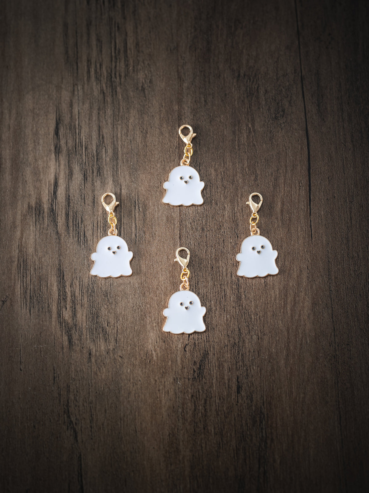 Four white enamel ghost charms with gold lobster clasps arranged on a dark wooden surface