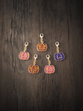 Five Halloween pumpkin enamel charms in pink, orange, brown and purple with gold lobster clasps on a dark wooden surface