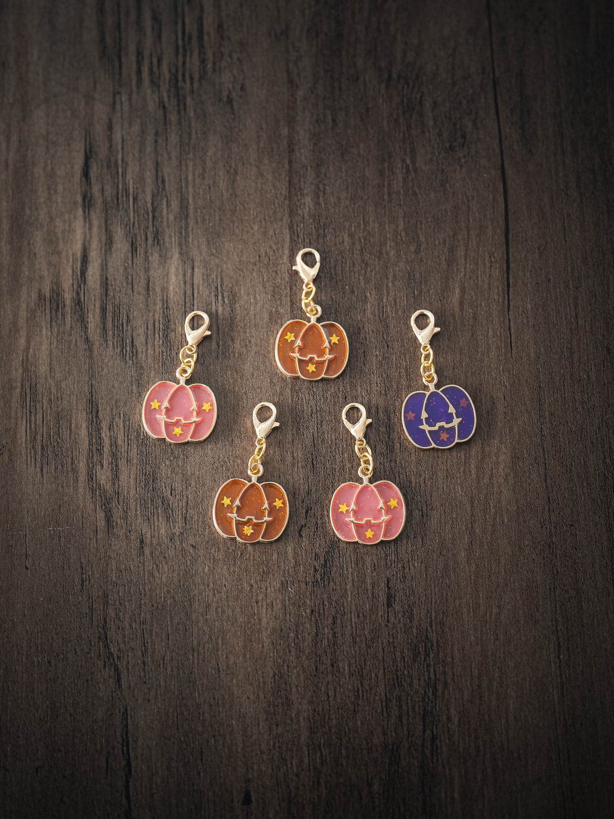 Five Halloween pumpkin enamel charms in pink, orange, brown and purple with gold lobster clasps on a dark wooden surface