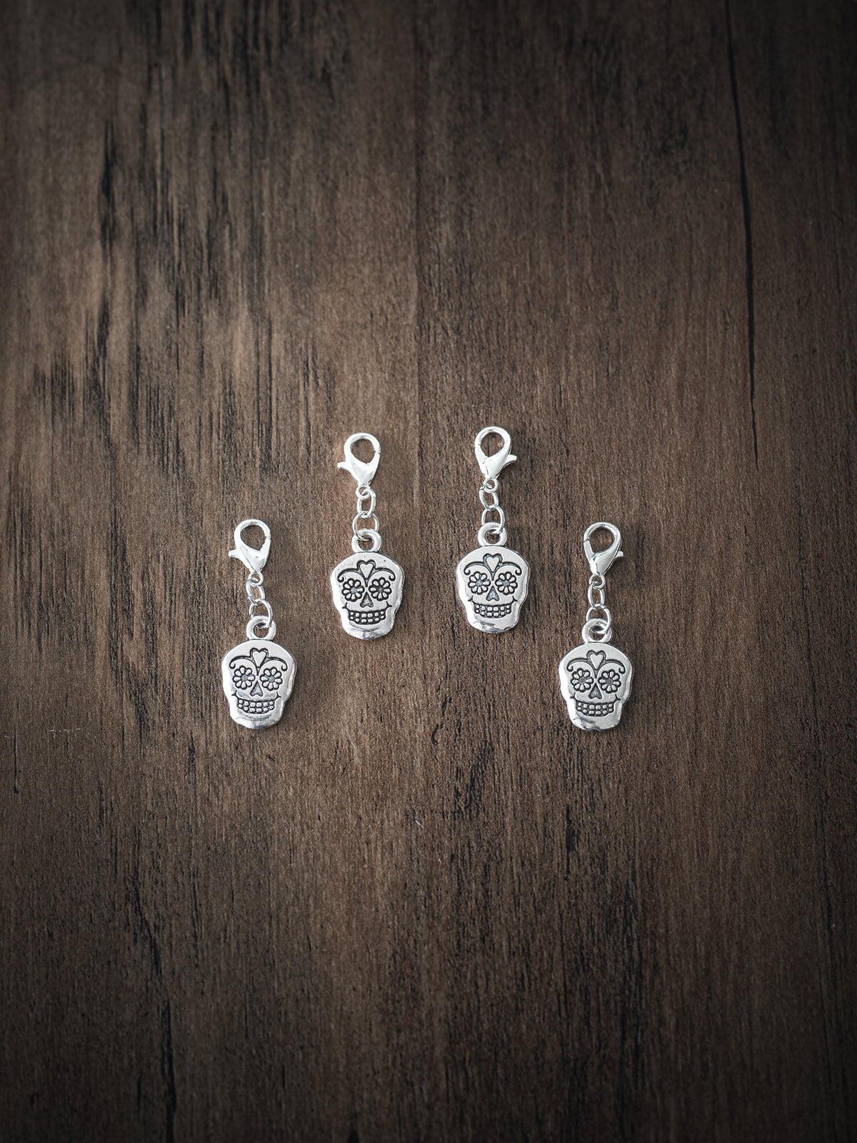 Four antique silver sugar skull charms with floral and heart face details and silver lobster clasps on a dark wooden surface
